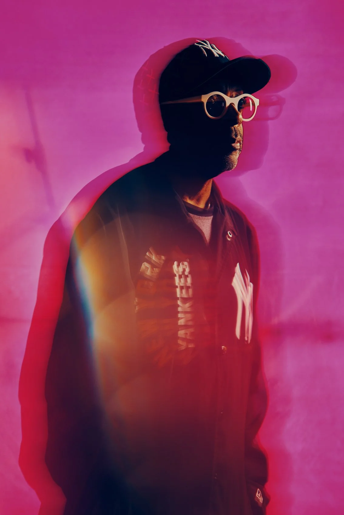 A man wearing a black New York Yankees cap, white sunglasses, and a black jacket with New York Yankees logo, standing against a pink background with a colorful light leak effect.