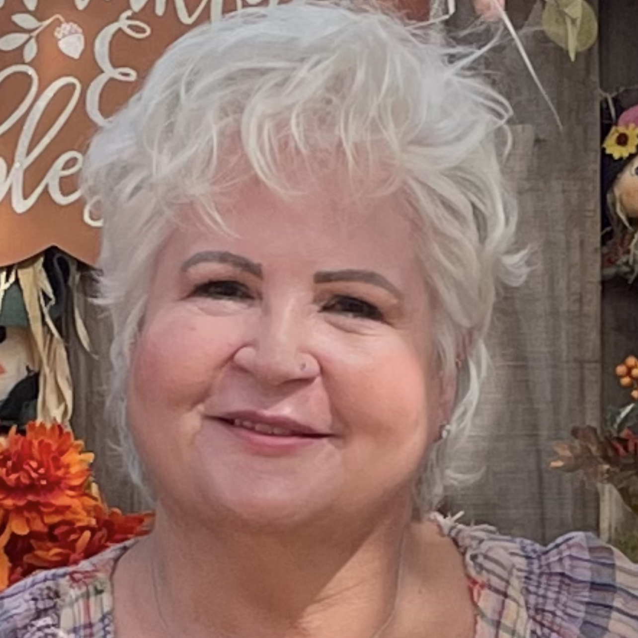 A smiling woman with short, curly, gray hair posing in front of a fall-themed background with orange flowers and decorative signs.