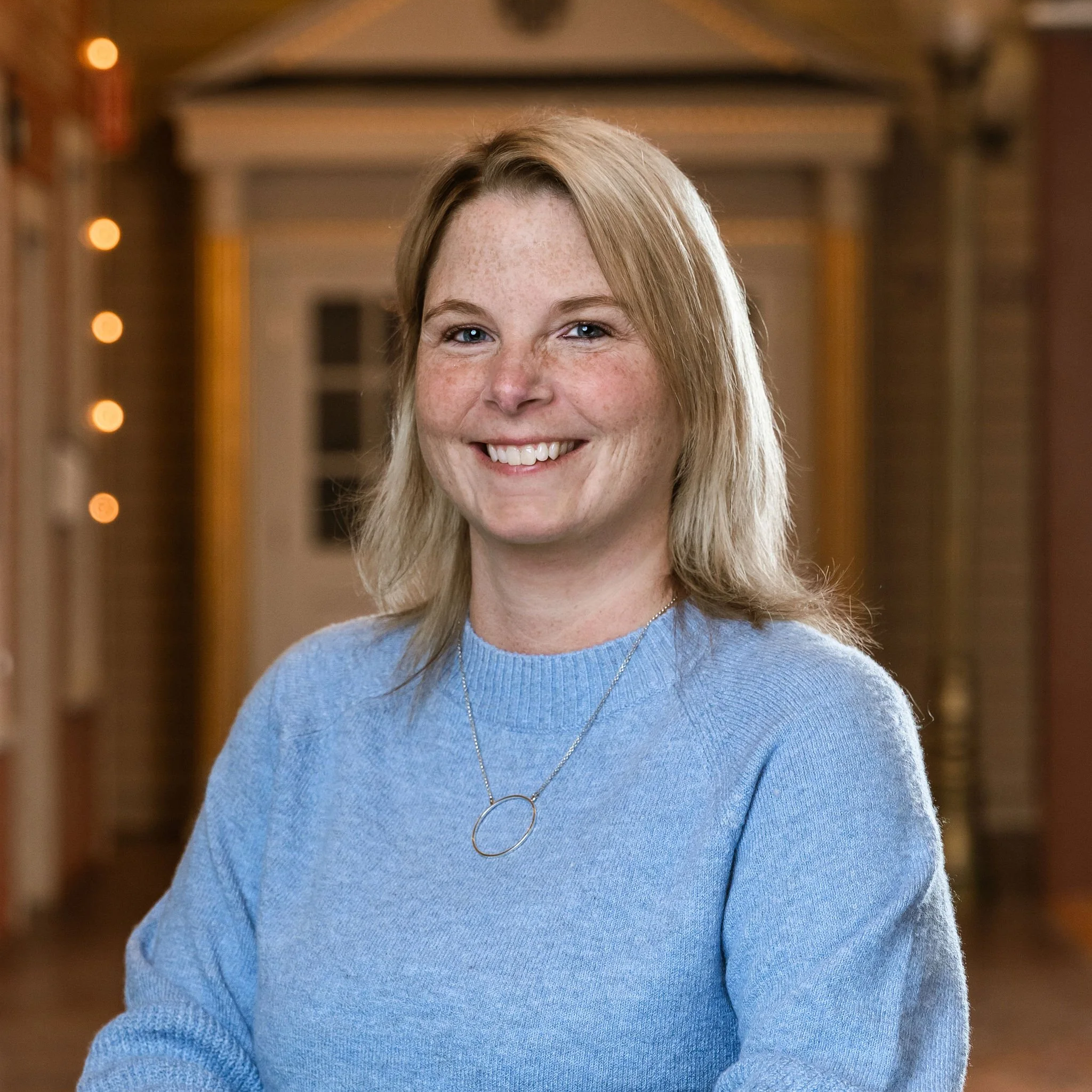 Close-up of a woman's face smiling, with shoulder-length blonde hair, wearing a light blue sweater and a silver necklace, in an indoor setting with warm lighting.
