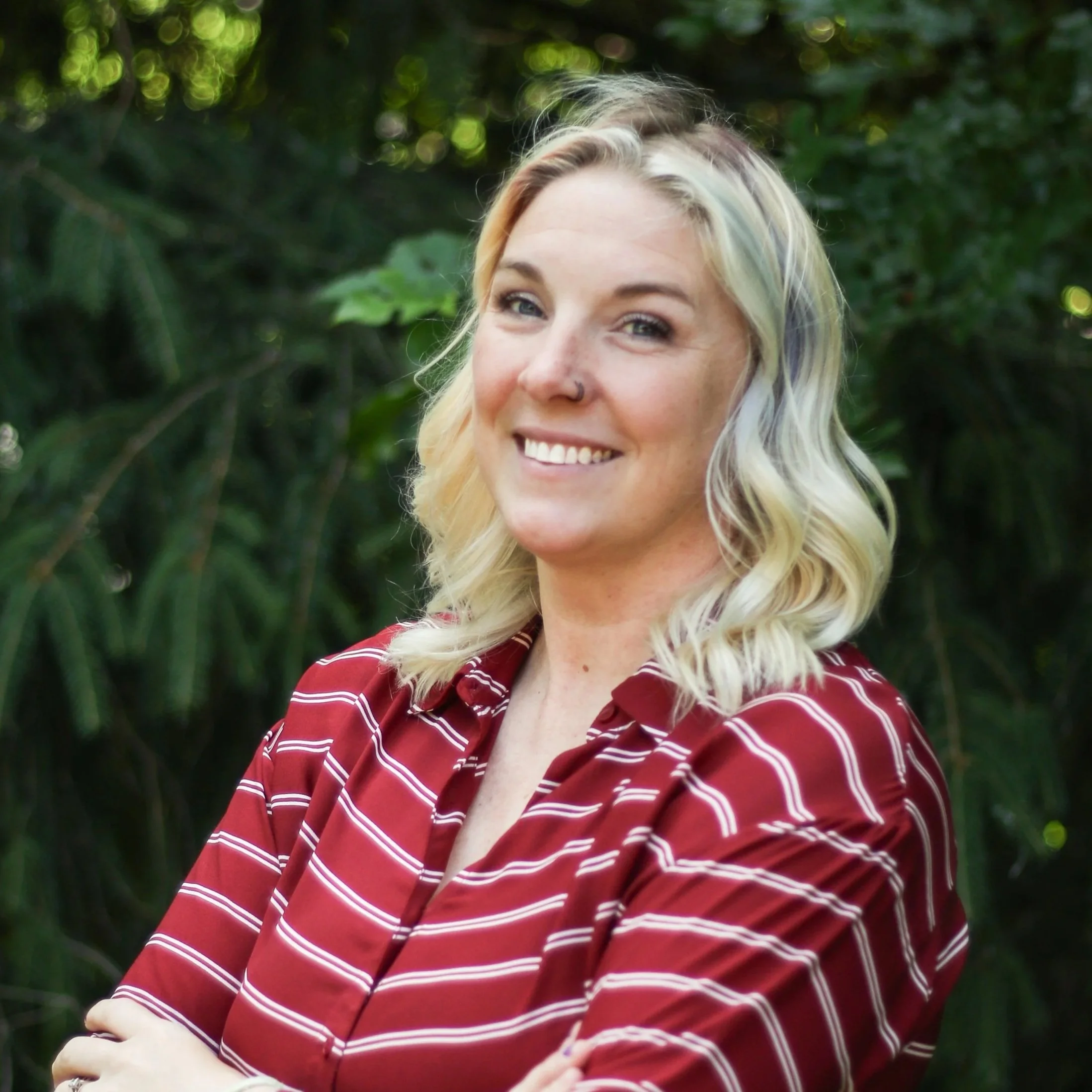 A woman with blonde hair, blue eyes, and a nose piercing smiling outdoors. She is wearing a red and white striped shirt and has a hand resting on her arm. The background features lush green foliage.