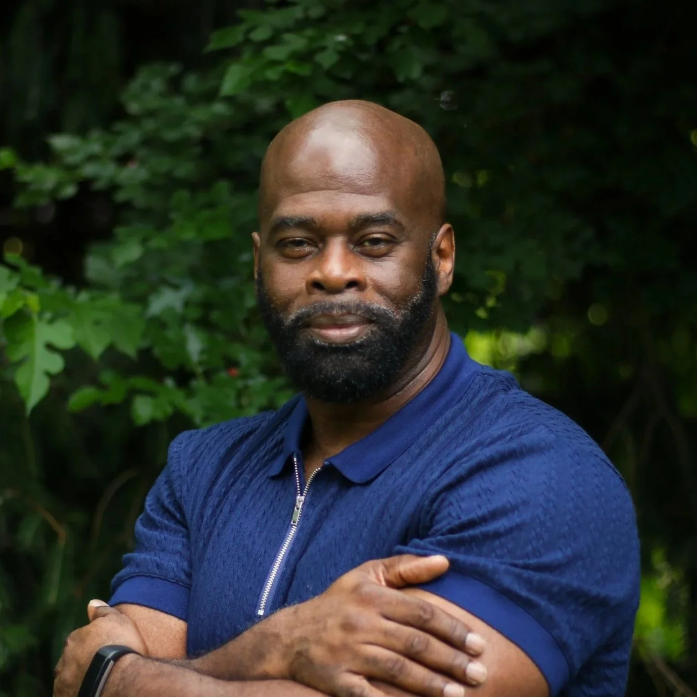 A man with a shaved head, beard, and crossed arms, standing outdoors in a lush green forest, wearing a blue zip-up jacket and a black smartwatch.