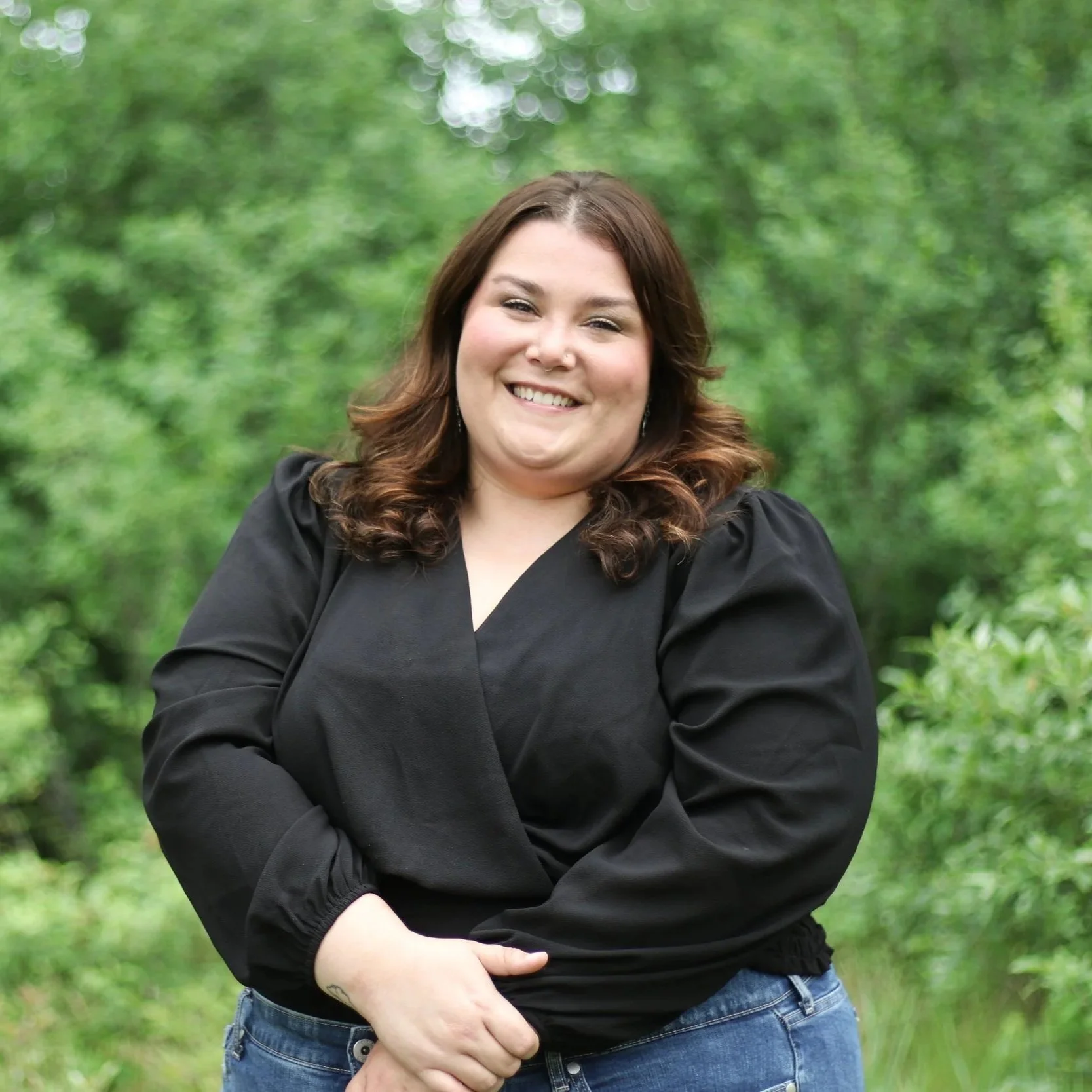 A woman with brown hair in loose waves, smiling, wearing a black top and blue jeans, standing outdoors in front of green trees.