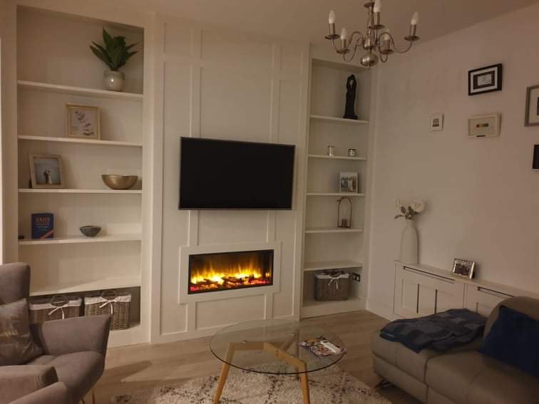 Living room with white built-in shelves, a wall-mounted flat-screen TV, and an electric fireplace below it. There is a gray armchair with a pillow on the left, a glass coffee table in front, and a gray sofa on the right. A white sideboard with a fram