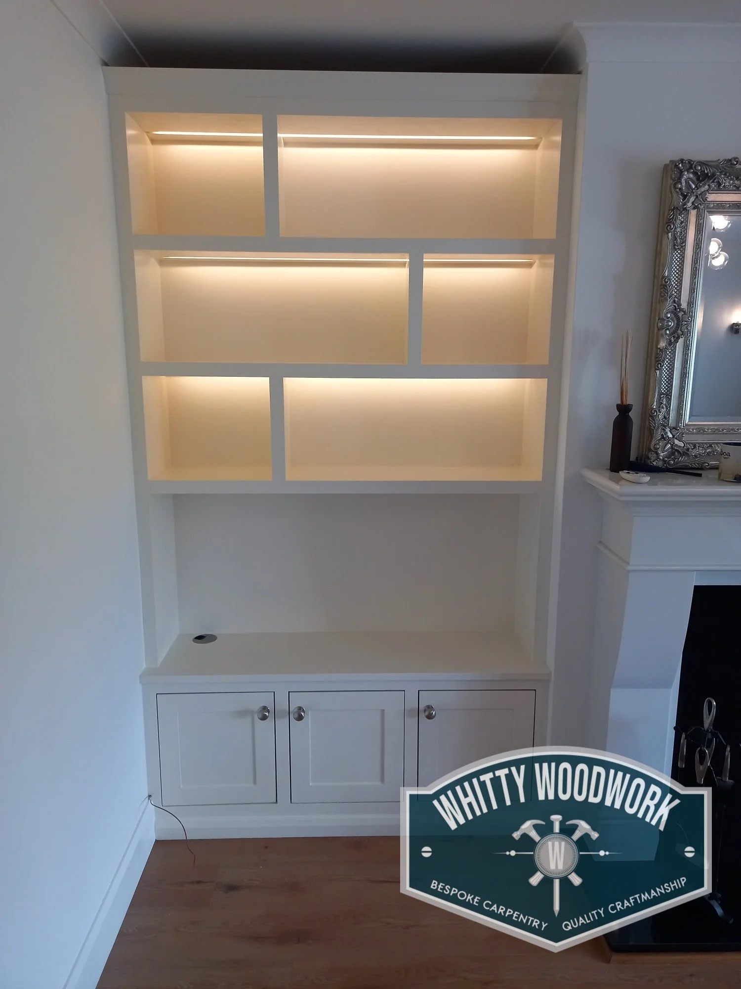 A white built-in cabinet with open shelves and cabinets below, situated against a wall in a room with hardwood flooring. Part of a large ornate mirror is visible to the right.