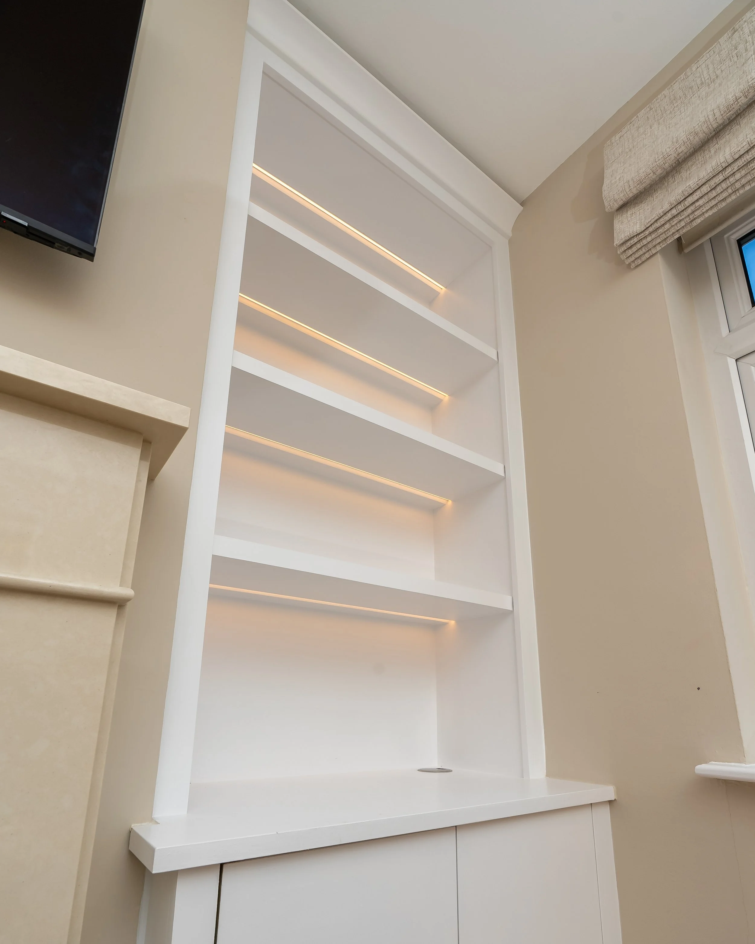 A white built-in bookshelf with five shelves with built-in lighting, situated in a corner near a window with beige Roman shades, in a room with beige walls.