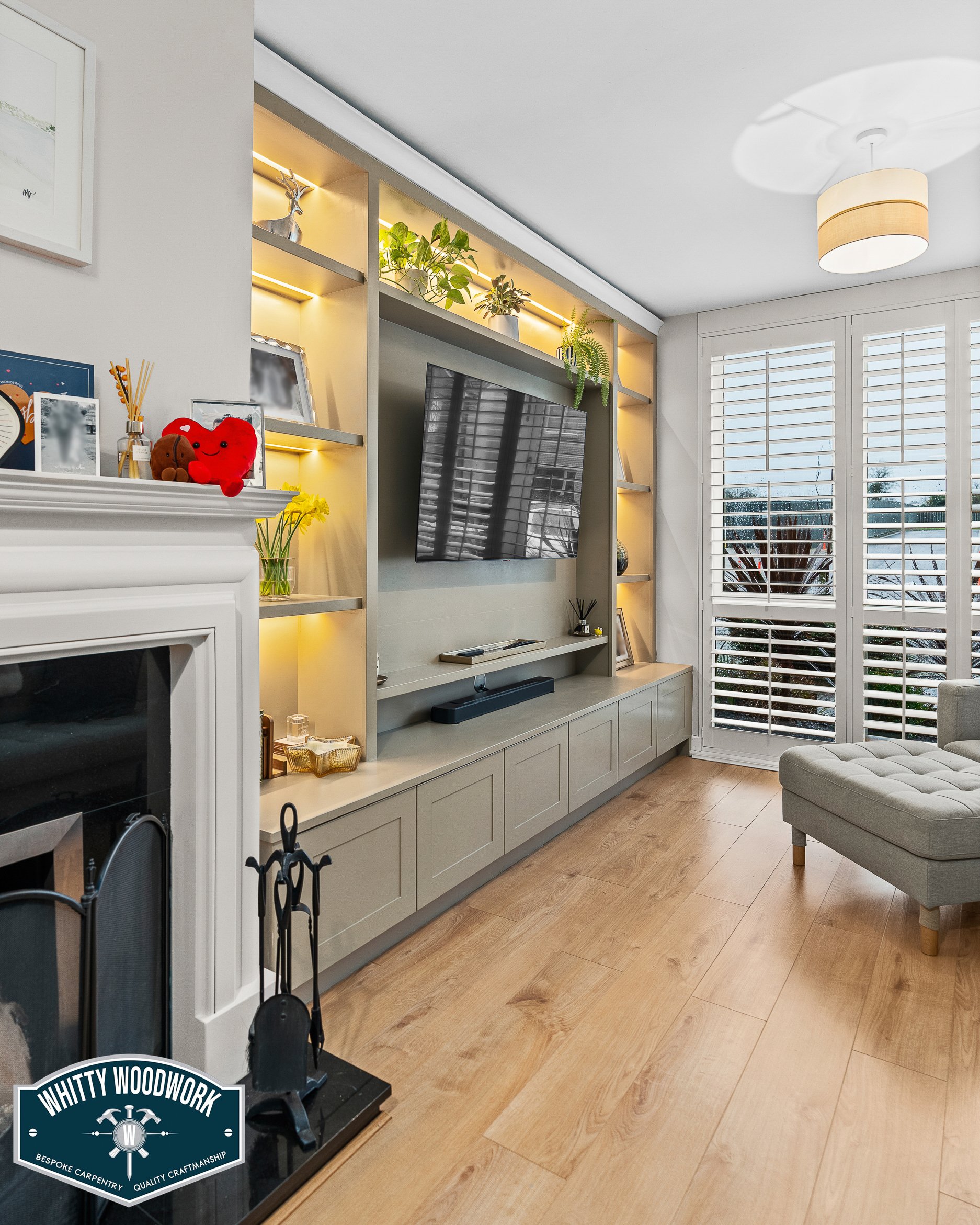 Living room with a wall-mounted flat-screen TV, window with slatted blinds, beige sofa, built-in shelving with plants, decorative items, and a fireplace mantel with a logo.