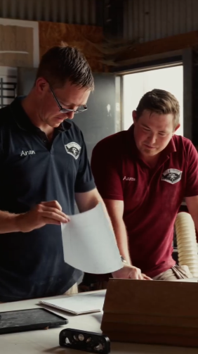 Two men in polo shirts reviewing documents at a table in a workshop or garage.