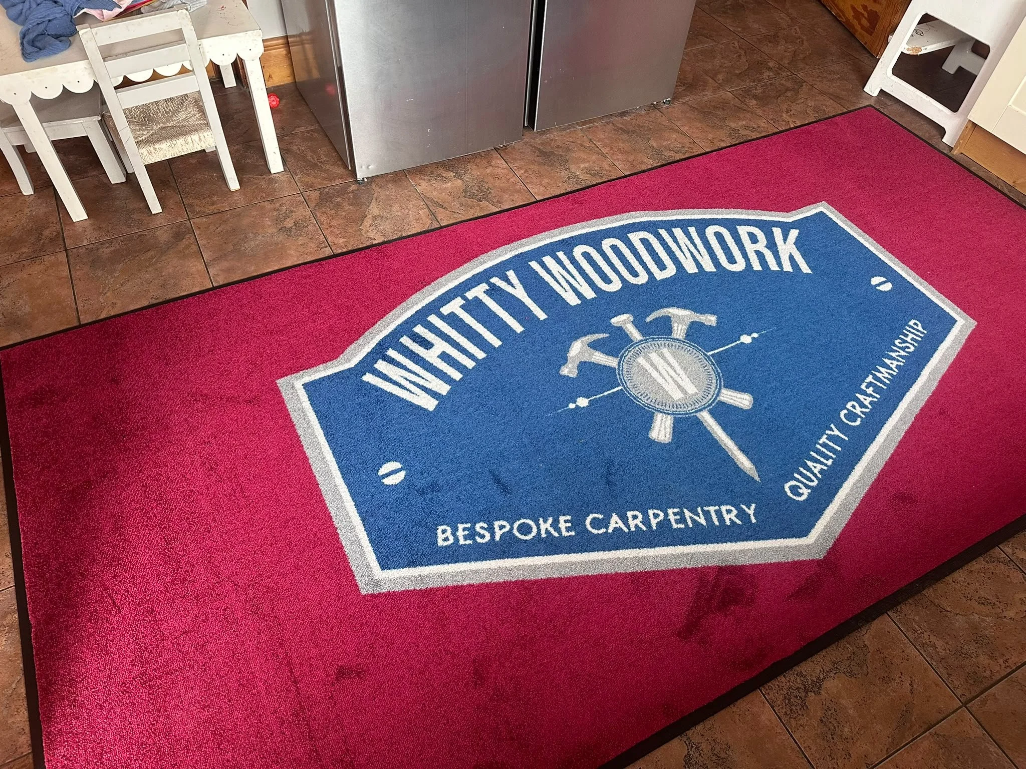 Close-up of a red doormat with a blue and white logo for a woodworking and carpentry business. Part of a dinette set and a stainless steel refrigerator are visible in the background.