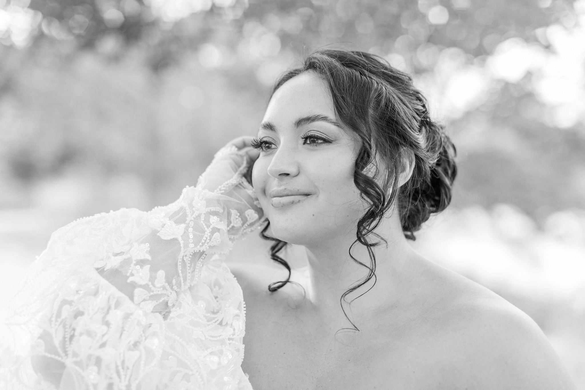 A woman with dark, curly hair styled in an updo, smiling gently, outdoors in natural light, with a blurred background of trees.