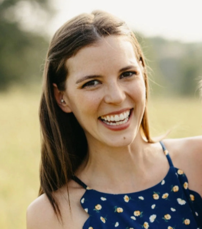 Close-up of a young woman with long brown hair smiling and showing her white teeth, against a light background.