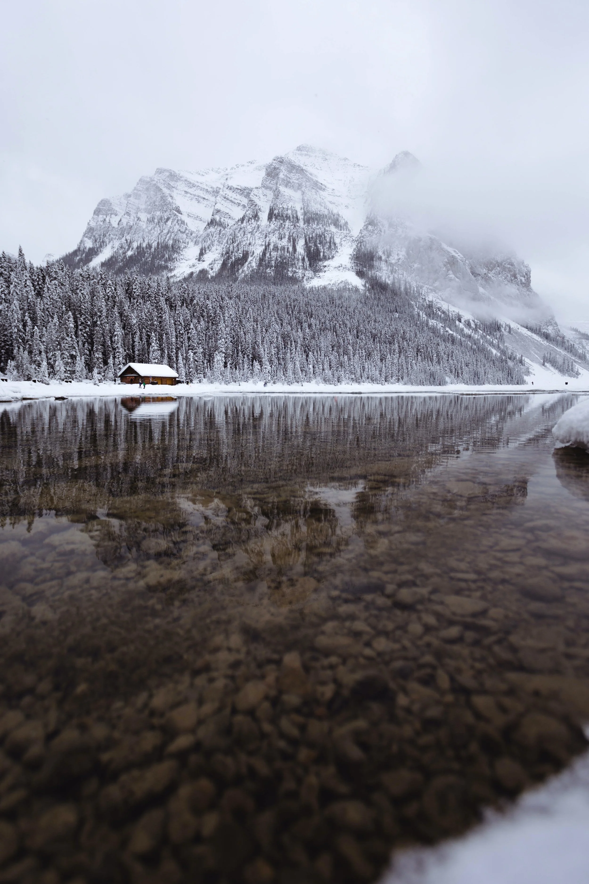 Cabin on the river at the base of a snowy mountain.