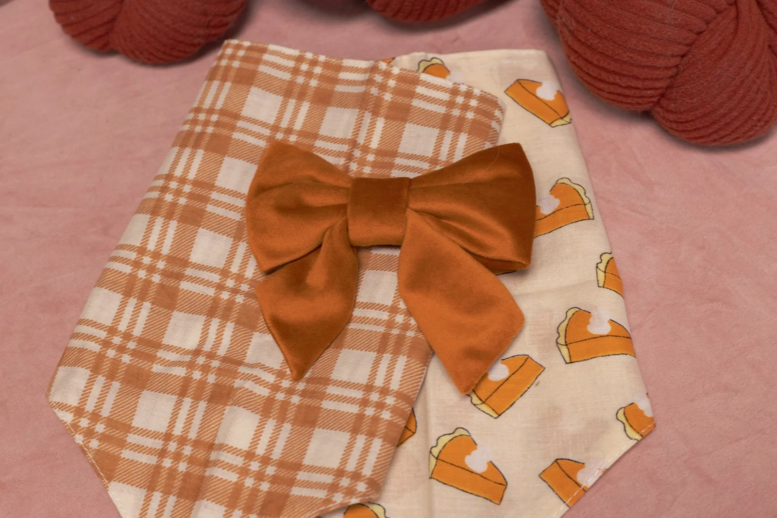 Two dog bandanas on a brown cushion. One has a pumpkin pattern, the other has a red and orange plaid design.