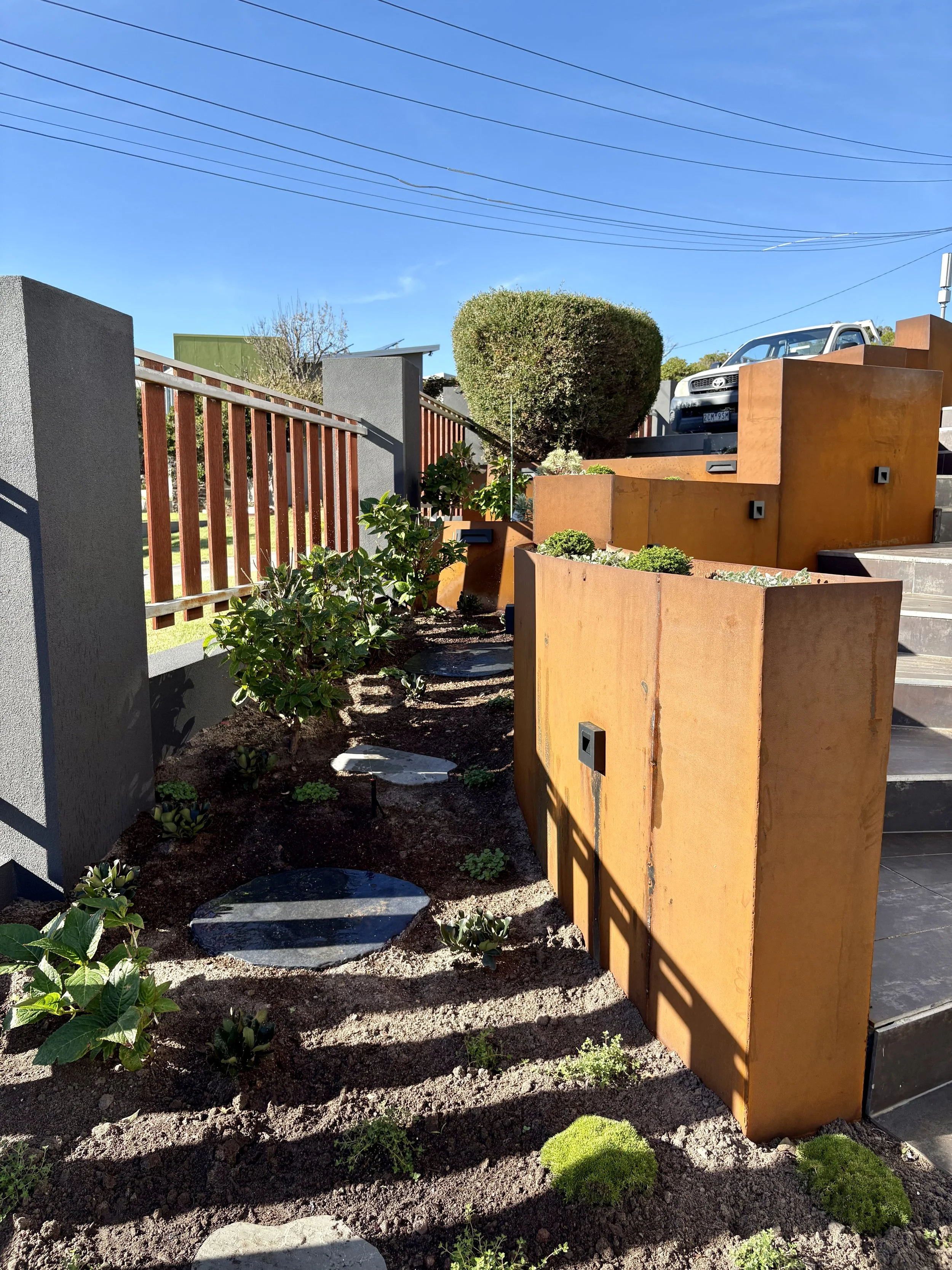 A landscaped garden with succulents, grasses, and trees, featuring a rust-colored metal wall and a stone pathway.
