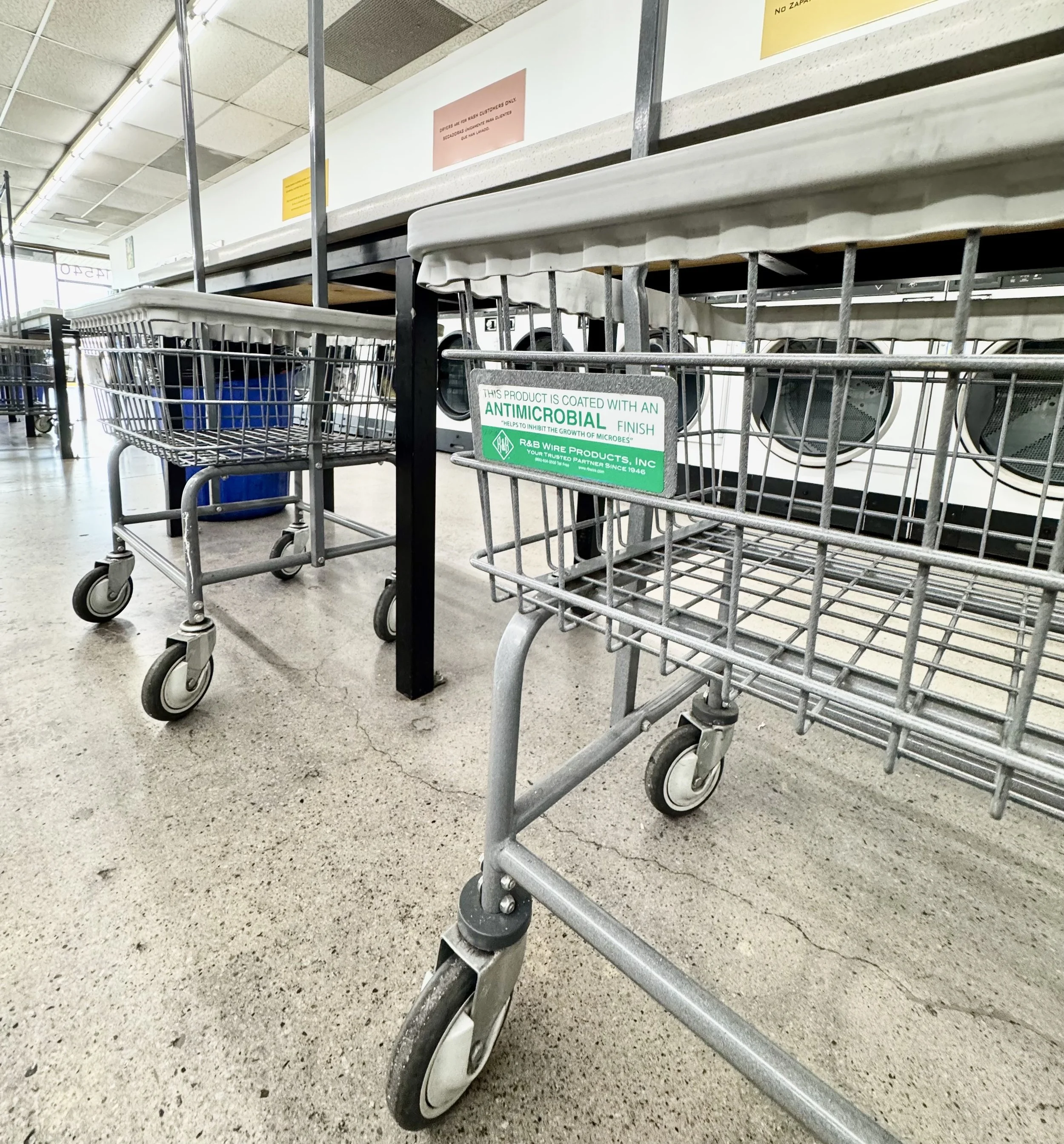 Empty shopping carts in a store aisle with a sign indicating antimicrobial coating, concrete floor, and laundry machines in the background.