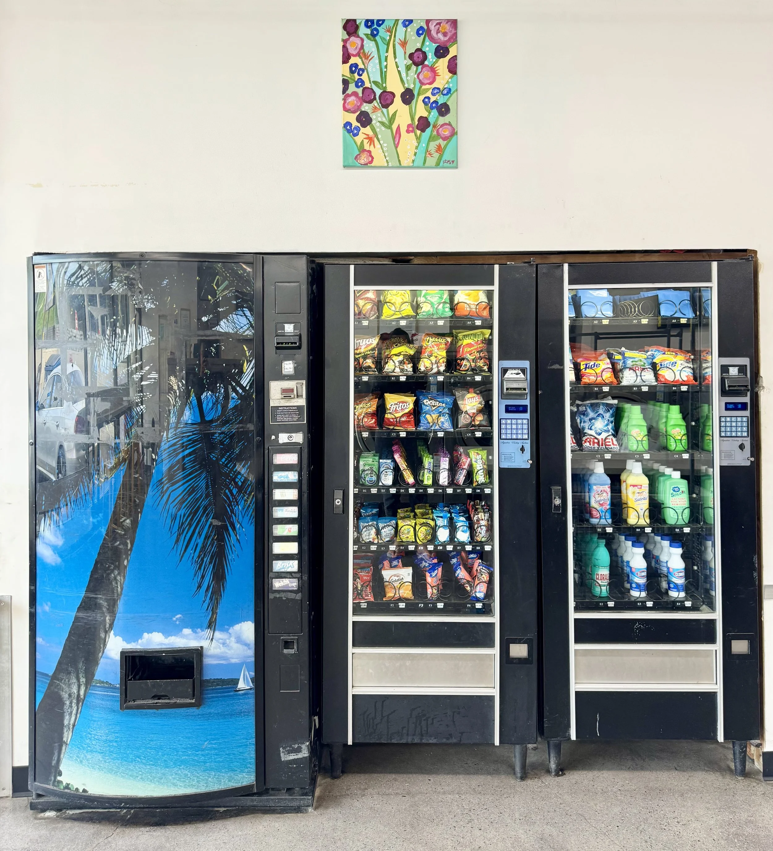 Two vending machines next to each other with snacks and drinks inside, and a colorful floral painting hanging above them on a white wall.