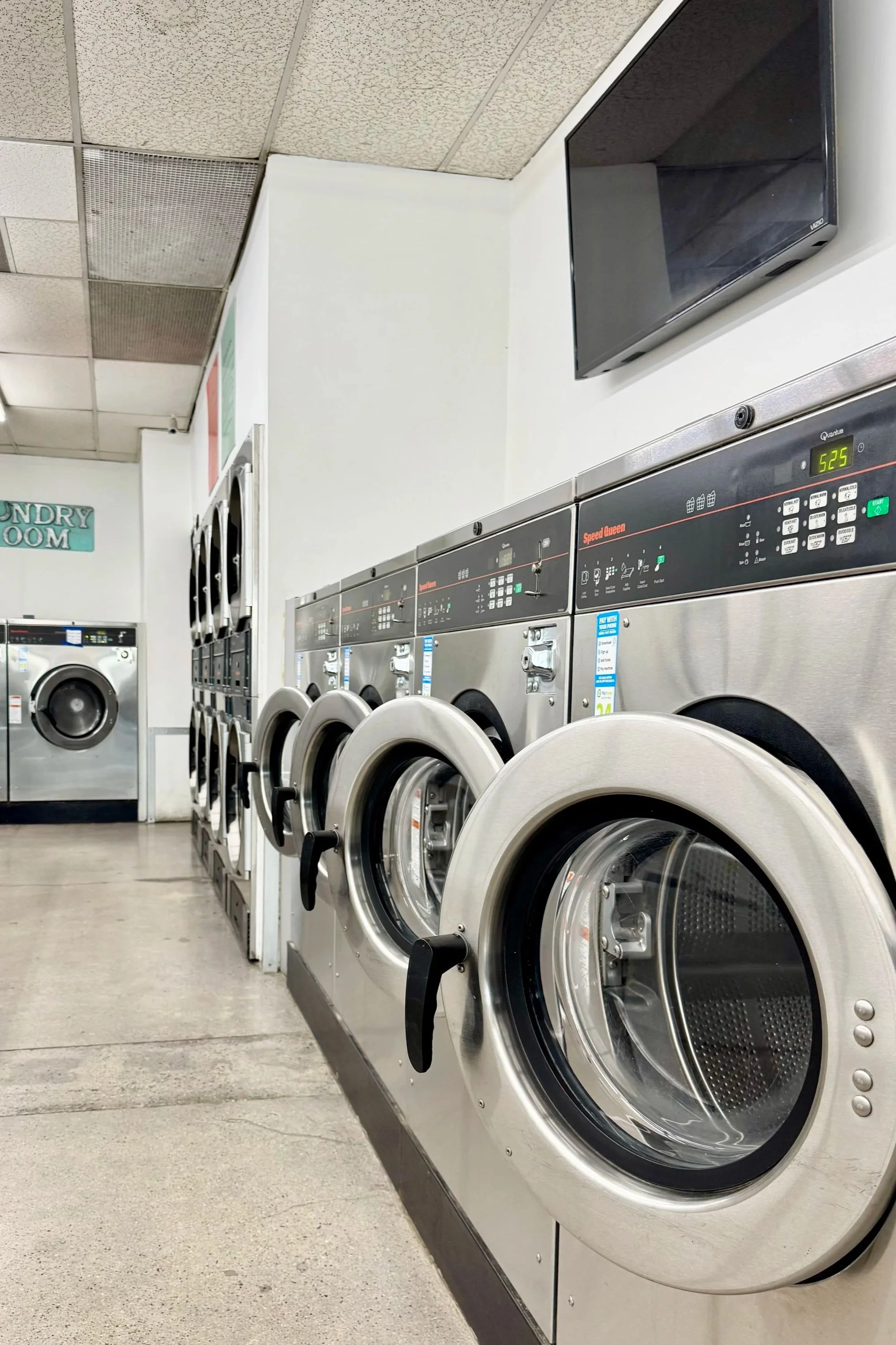 Interior of a laundromat with multiple silver washing machines lined up against the wall, a television mounted on the wall, and a sign reading 'Laundry Room' in the background.