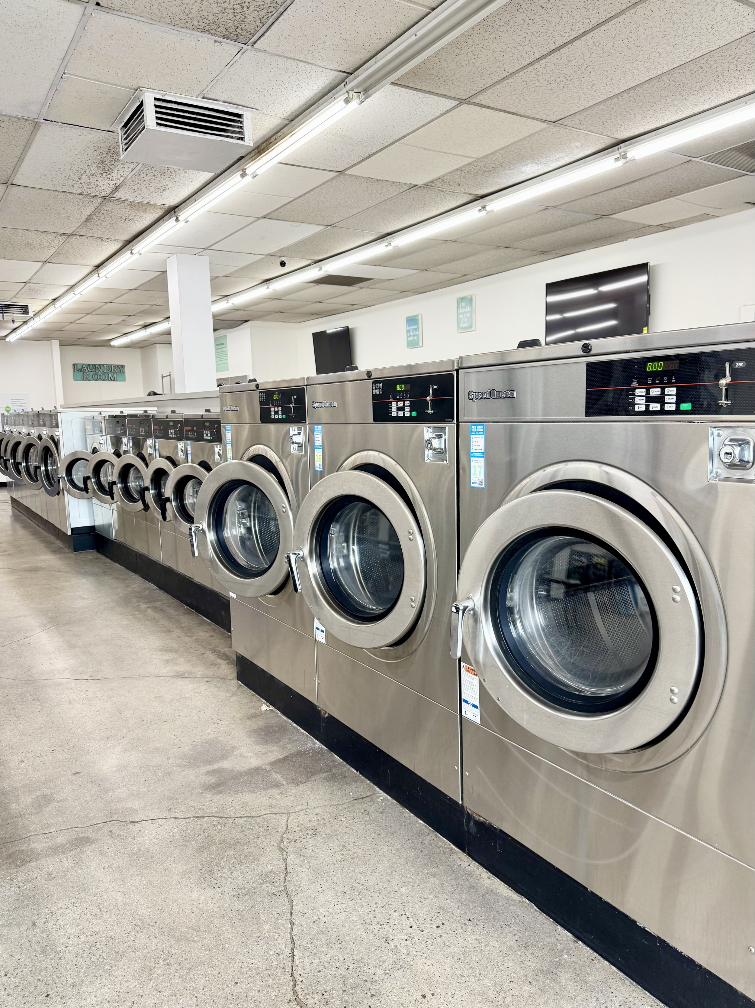 Multiple front-loading washers in a laundromat, with a sign that reads 'Laundry Room' visible in the background.