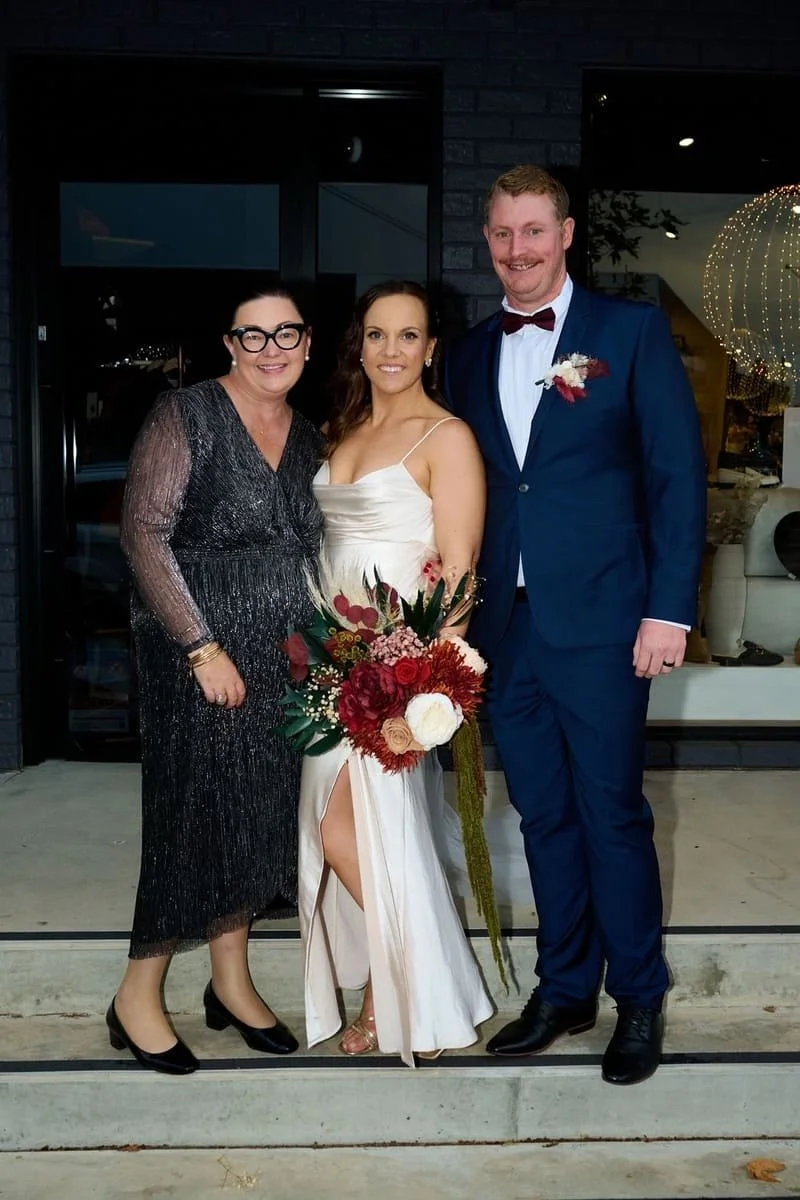 A woman in a white wedding dress holding a bouquet of red, white, and pink flowers, standing between a woman in a sparkling black dress and a man in a blue suit with a red bow tie, outside a building with dark brick walls at nighttime.