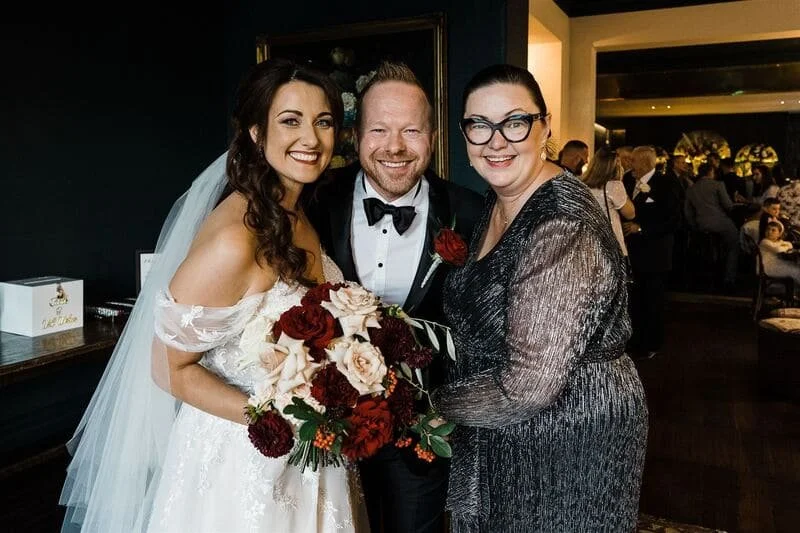 Three people at a wedding reception: a bride in a white gown holding a bouquet, a groom in a tuxedo, and a woman in a metallic dress with glasses, smiling and posing together.
