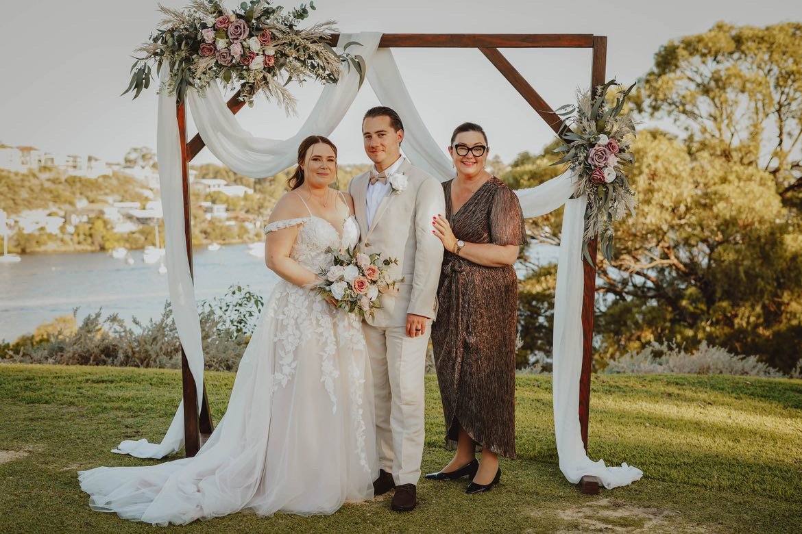 A bride in a white wedding gown with floral embroidery holding a bouquet, a groom in a white suit with a bow tie, and a woman in a dark patterned dress standing under an outdoor wedding arch decorated with flowers and white fabric, with a water and trees in the background.