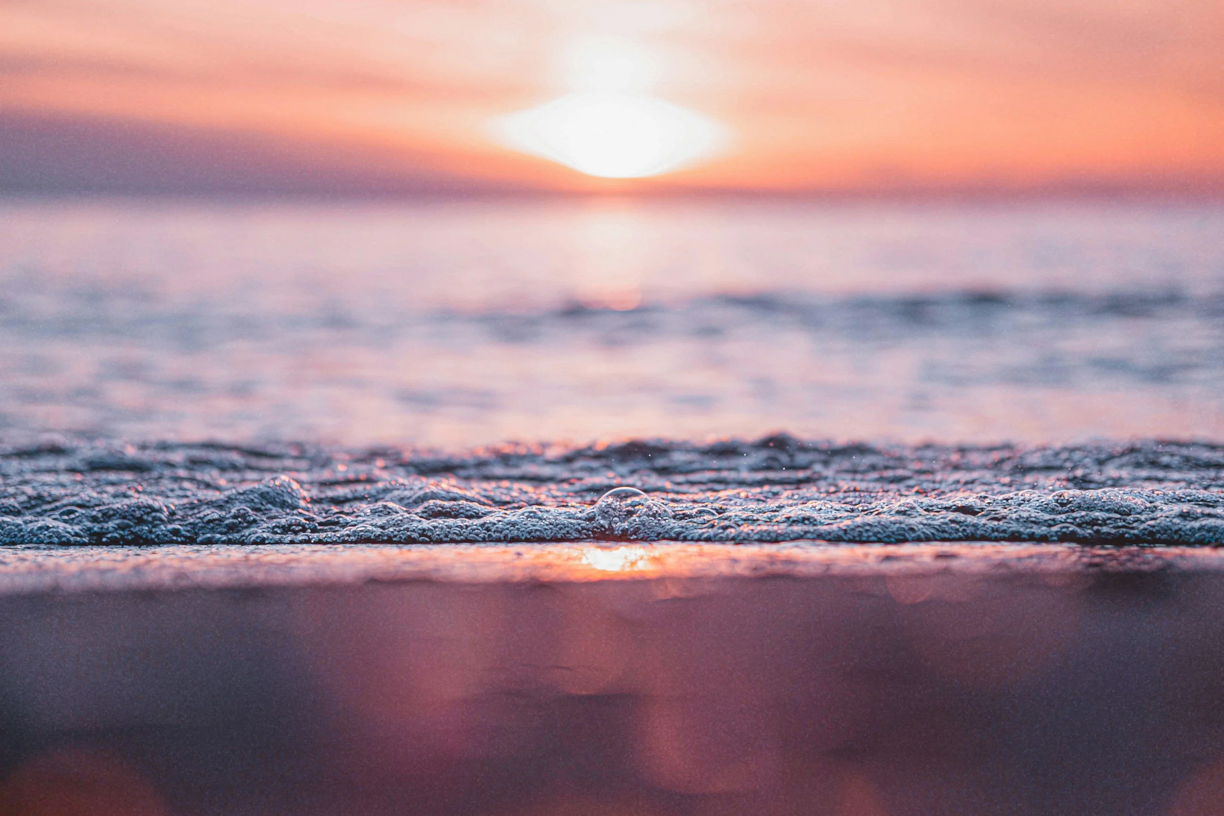 A serene beach scene during sunset with pink and orange hues in the sky, gentle waves lapping the shore, and foam on the water.