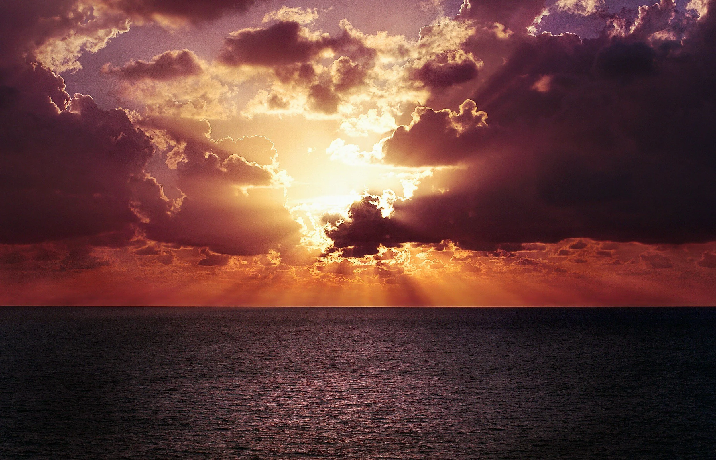 A sunset over the ocean with dark clouds partly covering the sky and the sun shining through, creating a glow and reflections on the water.