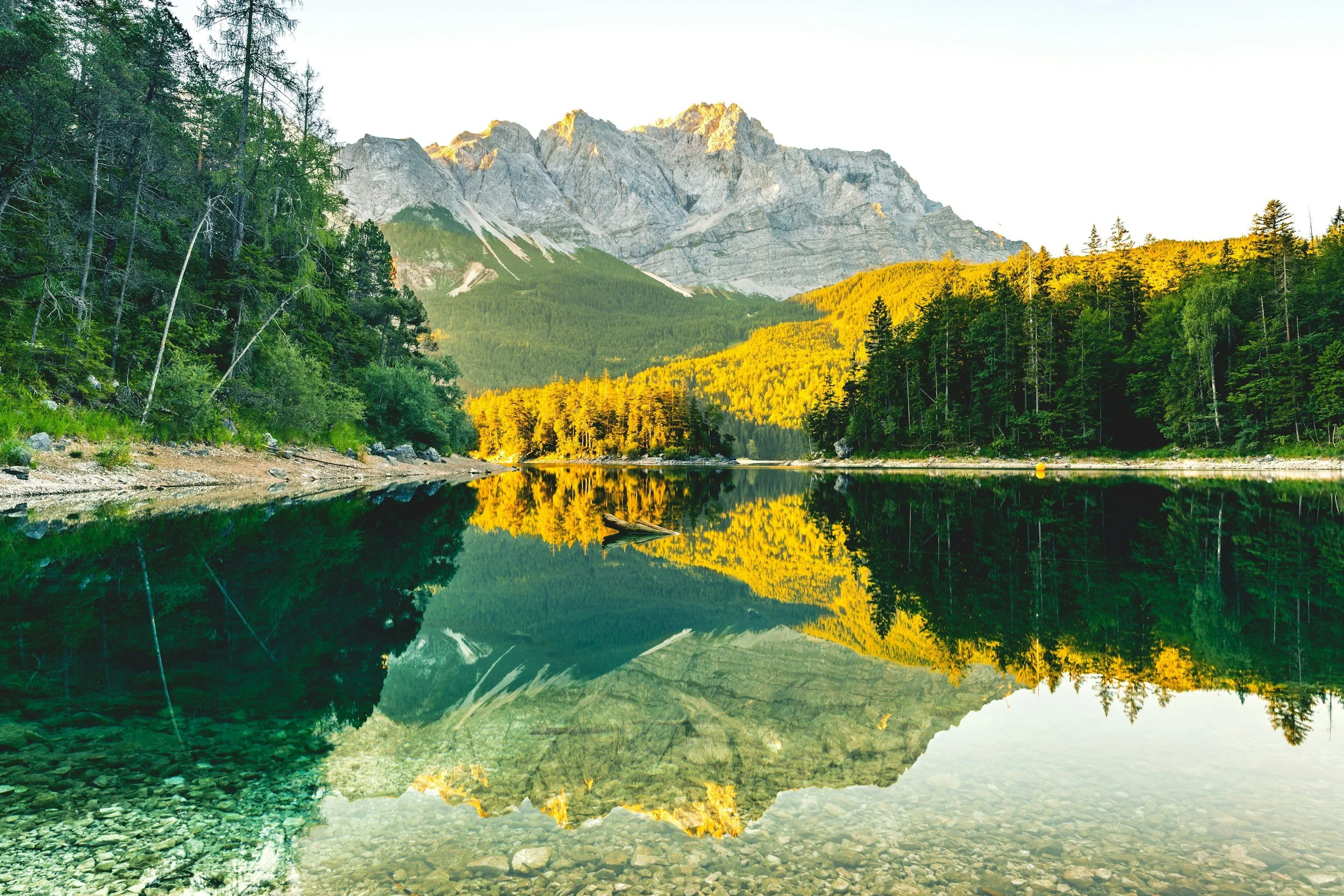 Scenic view of a mountain with rocky peaks, surrounded by green forest, reflected in a calm lake in the foreground.