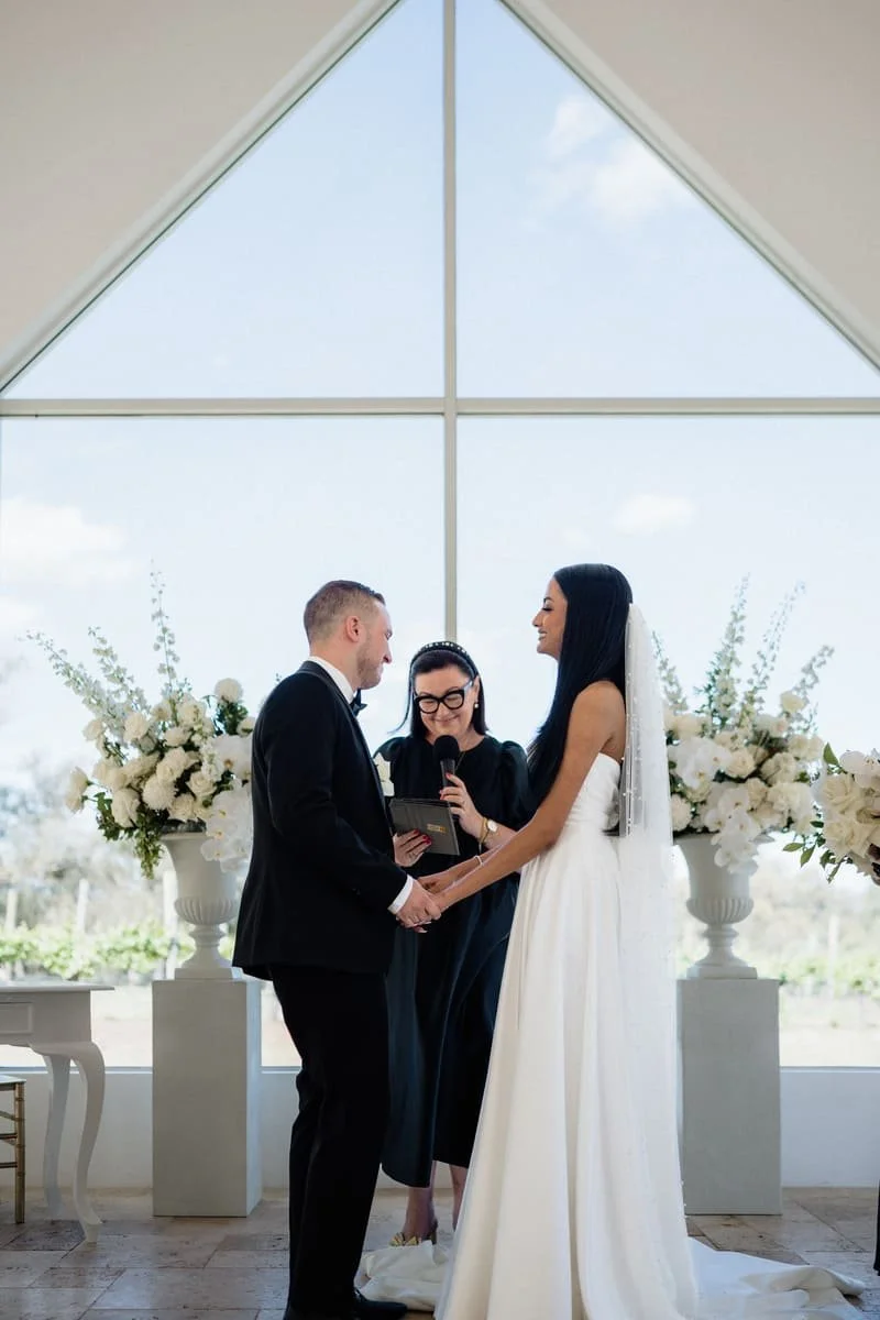 A couple getting married and exchanging vows in front of a large window with floral arrangements on either side during a wedding ceremony.