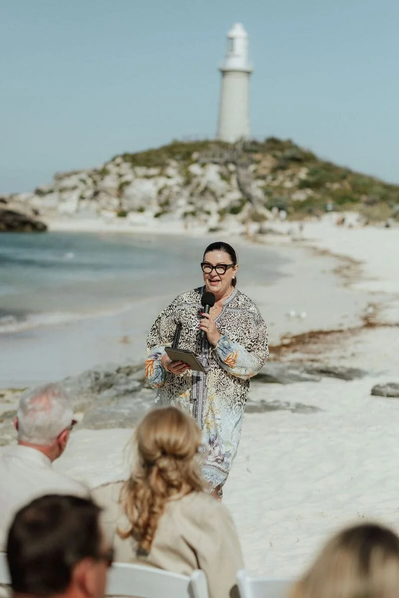 A woman holding a microphone and a tablet, speaking to an audience on a beach with a lighthouse in the background.