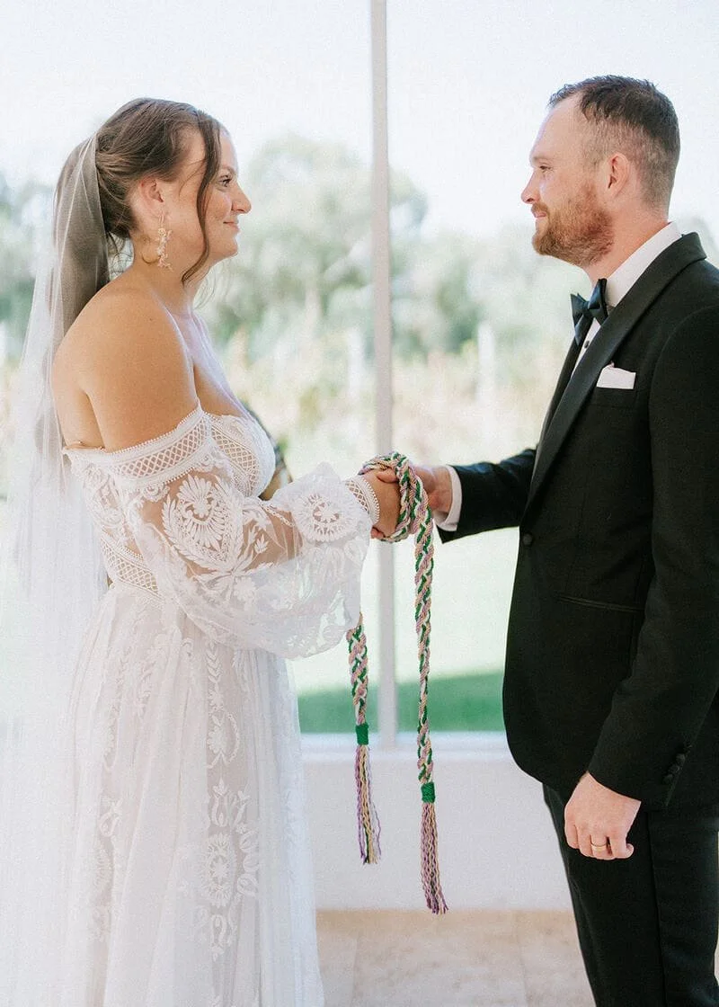 Bride and groom holding hands during their wedding ceremony indoors, with a window in the background.