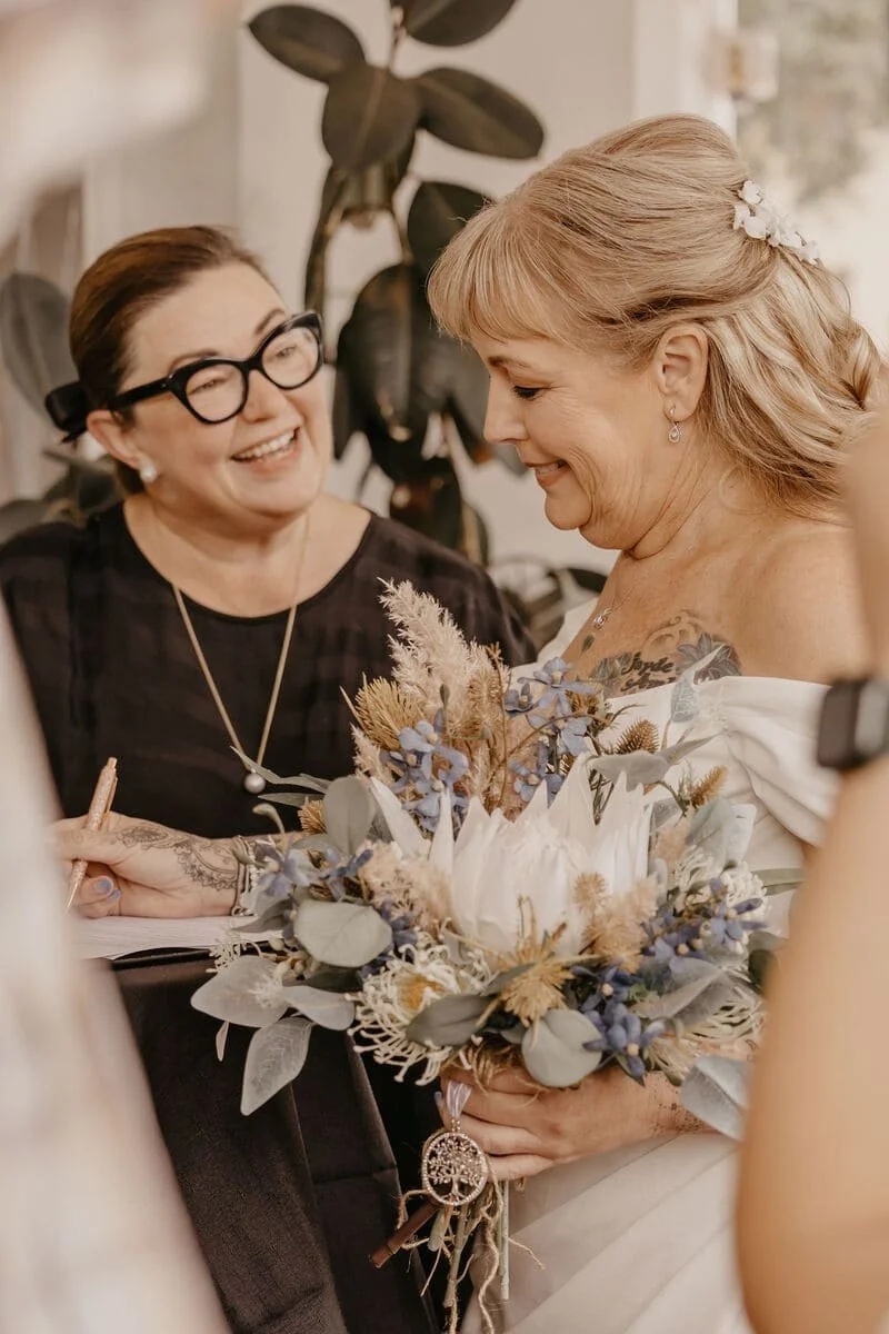 A woman in a white wedding dress holding a bouquet of dried and fresh flowers, smiling at another woman, who is signing a document and laughing.