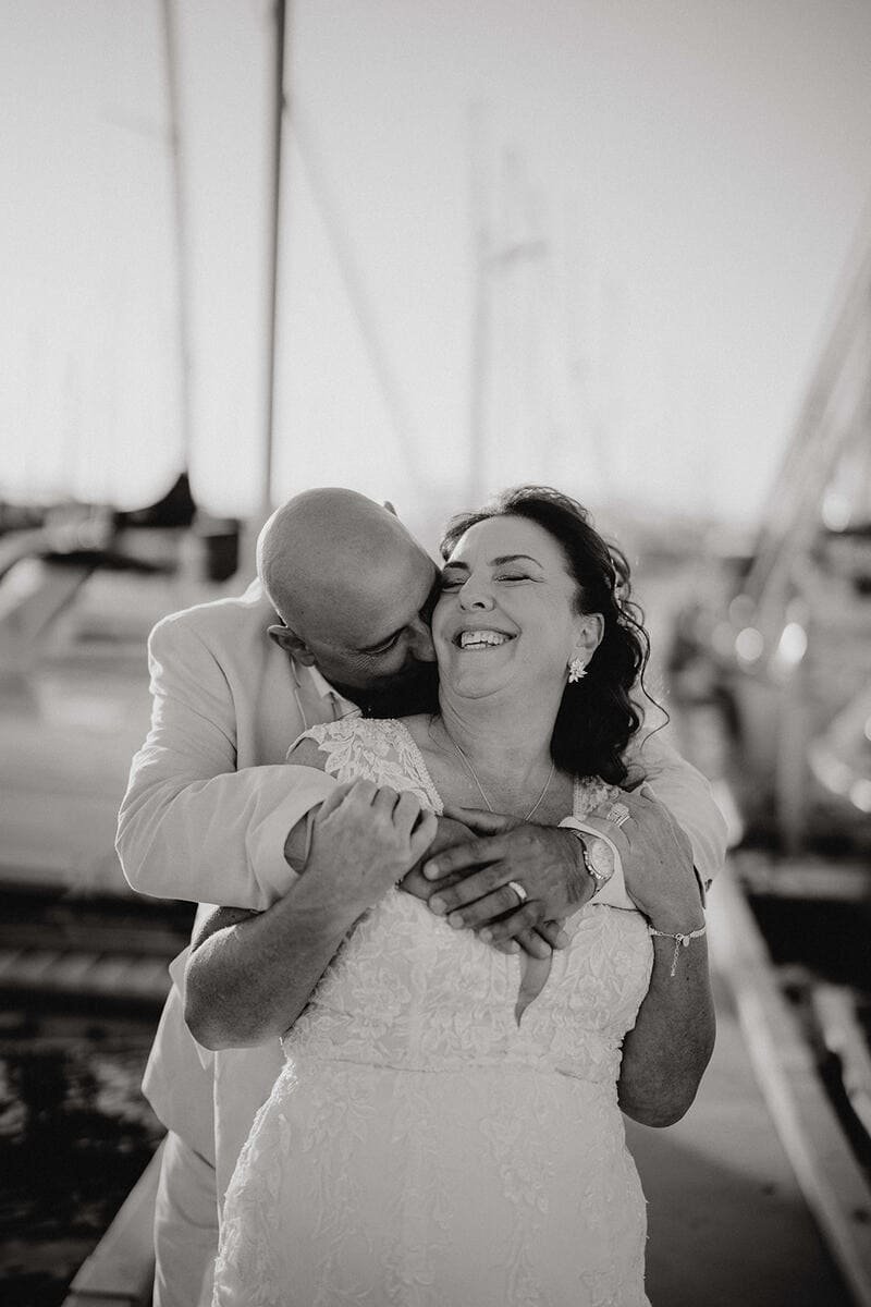 A joyful woman in a wedding dress being embraced from behind by a man in a suit, both smiling, at a boat dock.