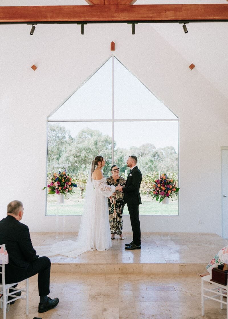 A wedding ceremony with a bride and groom holding hands and exchanging vows in front of an officiant in a room with large window and floral arrangements.