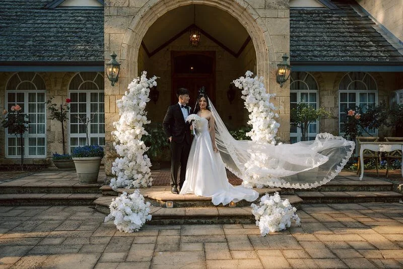 Bride and groom standing together on wedding steps, surrounded by white floral arrangements, in front of a stone building with arched entrance during sunset.
