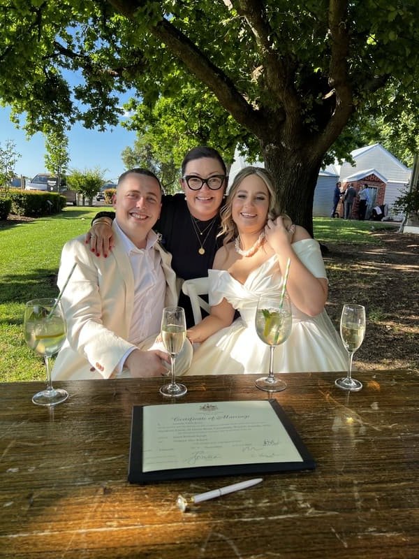 Three people, two women and one man, sitting together outdoors under a large tree with green leaves. They are smiling and dressed nicely, with the woman on the right wearing a white dress. There are four glasses of champagne on the table in front of them and a document at the center of the table. The background shows a clear blue sky and a few buildings.