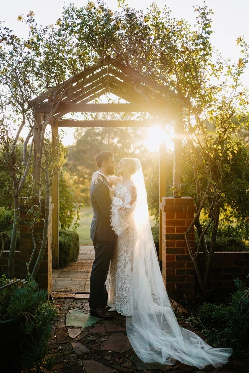 A bride and groom share a moment under a wooden archway at sunset, surrounded by trees and greenery, on their wedding day.