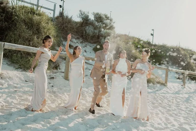 A group of four women and one man in formal attire laughing and enjoying themselves on a sandy beach with a wooden fence and greenery in the background.