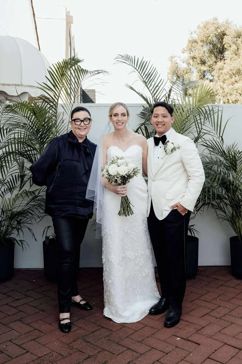 A bride in a white wedding dress holding a bouquet, standing between a woman in black and a man in a white tuxedo jacket outside, with potted plants and trees in the background.
