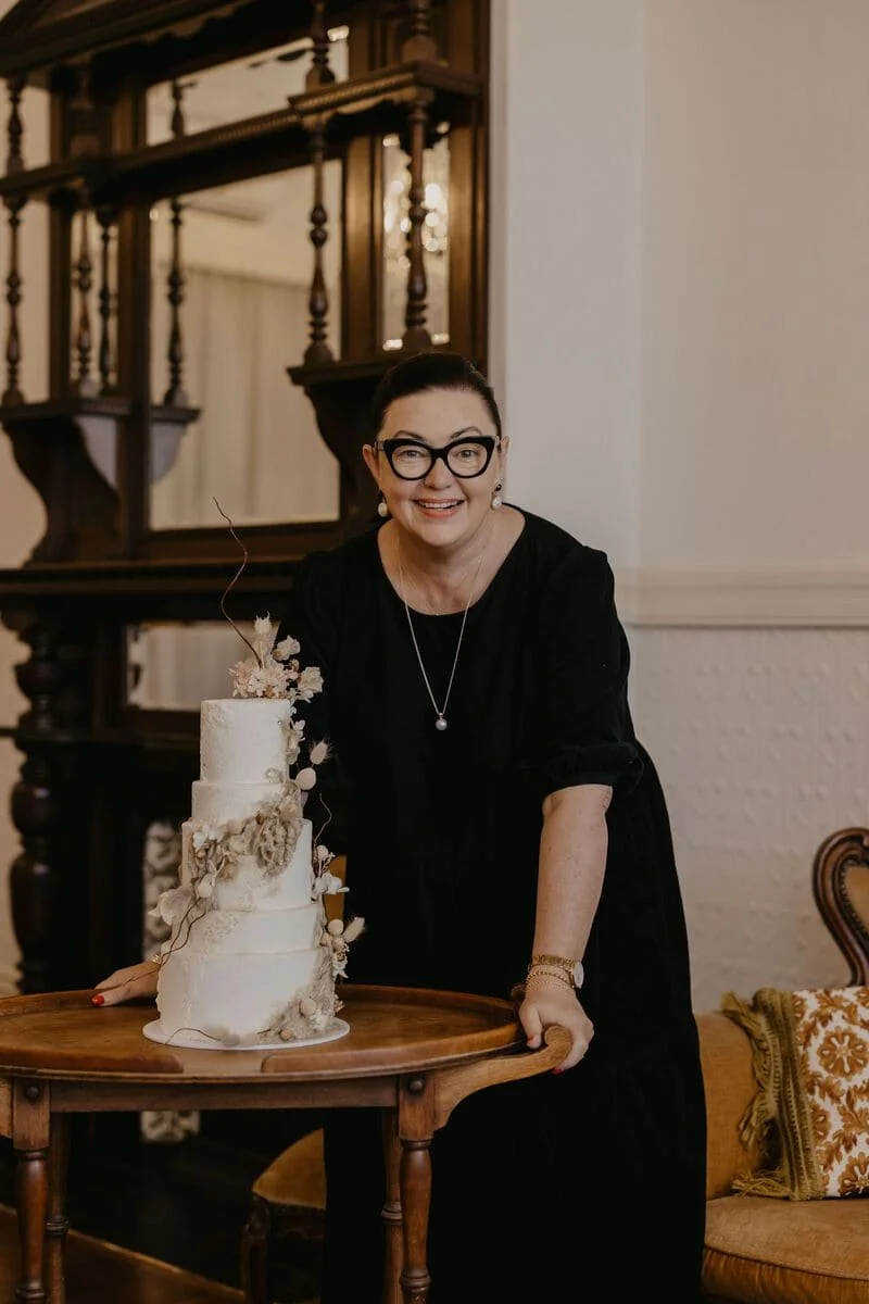 A woman with black hair, glasses, and earrings smiling and leaning on a small round wooden table with a three-tiered white wedding cake decorated with flowers and dried leaves, in an elegant, classic room.