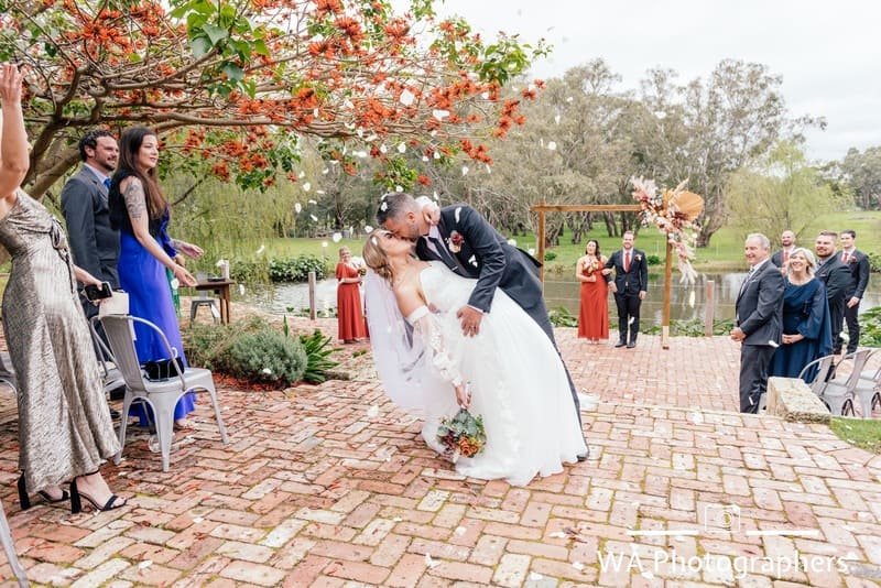 A newlywed couple sharing a dip and kiss during outdoor wedding ceremony, surrounded by guests, brick pathway, trees, and river background.