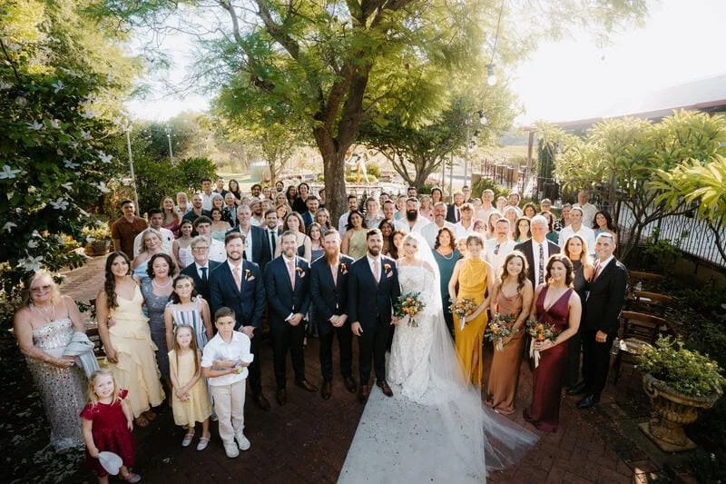 A large group of people gathered outdoors for a wedding, including a bride and groom, family, and friends, with trees and sunlight in the background.