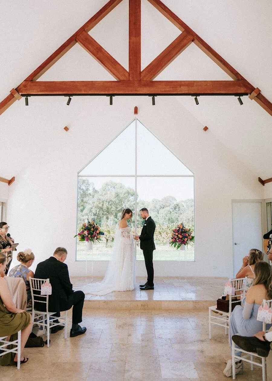 A wedding ceremony indoors with a bride and groom holding hands at the altar, in front of a large window with trees outside. Guests are seated on either side, and floral arrangements are present.