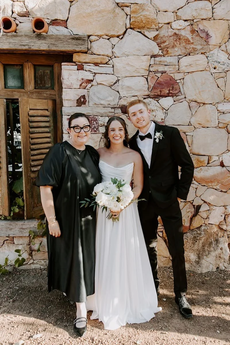 A woman in a black dress, a bride in a white wedding gown holding a bouquet, and a groom in a black tuxedo with a bow tie standing together in front of a stone wall, smiling.