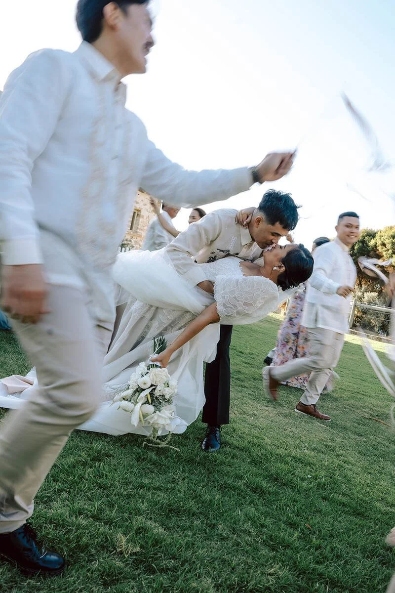 A couple is kissing at a wedding celebration, with the groom dipping the bride while holding a bouquet of flowers. Guests are around them, some holding hats and others smiling. The scene takes place outdoors on a grassy area under a clear sky.