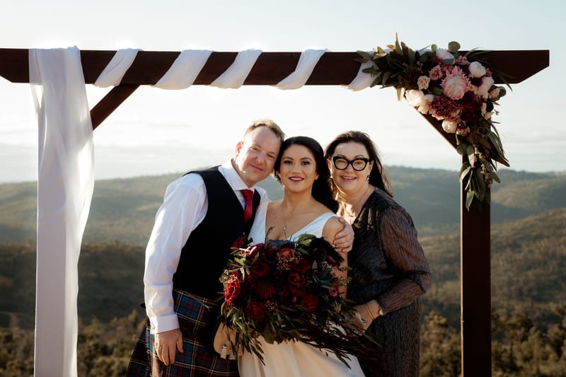 Three people pose under a wooden wedding arch decorated with pink and white flowers and white fabric ribbon on a hillside with a scenic landscape in the background.