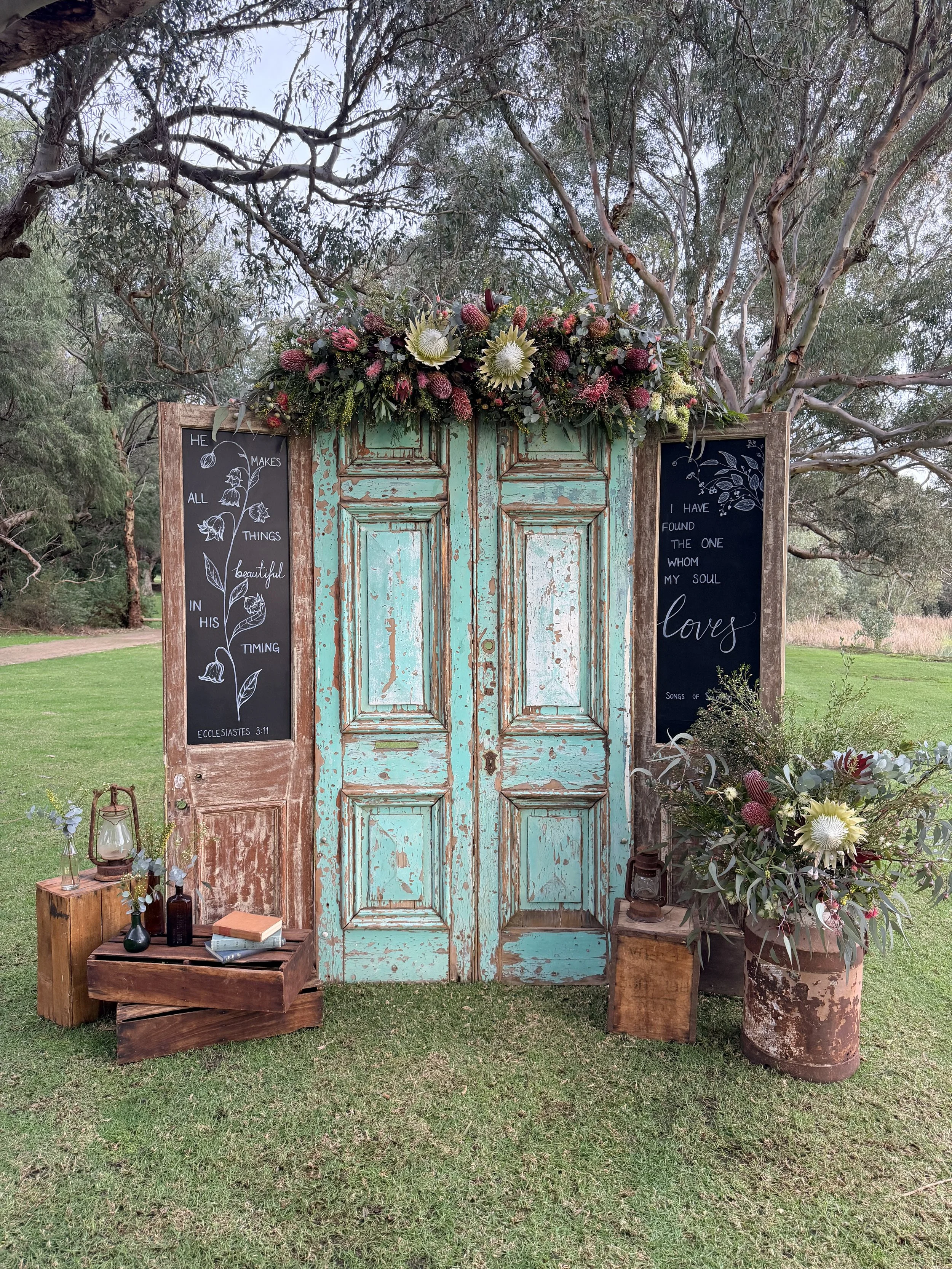 Rustic outdoor decoration featuring a weathered turquoise double door with floral arrangements on top, flanked by blackboards with quotes, and sitting on grassy ground with trees in the background.