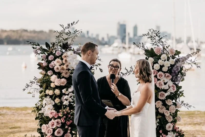A couple getting married outdoors near a body of water, with a wedding officiant holding a microphone and plants with pink and white flowers in the background.