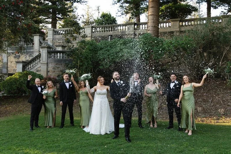A wedding celebration outdoors with a group of people dressed in formal attire, some holding bouquets, standing on a green lawn with trees and a stone staircase in the background. They are celebrating and smiling, with champagne and confetti in the air.
