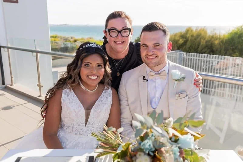 A smiling bride and groom sitting at a table with a woman standing behind them, on a balcony with ocean views in the background. The bride wears a white lace wedding dress and a tiara, the groom wears a white suit with a bow tie, and the woman behind them has glasses and a black top.