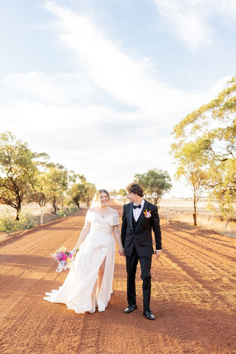 A bride and groom walking hand in hand on a dirt road with trees on either side during a sunny day.
